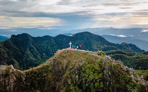 A group of hikers standing near a white cross on the rocky summit of Volcán Barú overlooking layered green mountain ranges